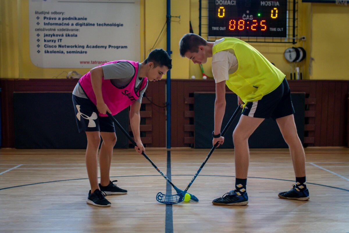 Two teenagers face off in a floorball match inside a sports gym with equipment visible in the background.