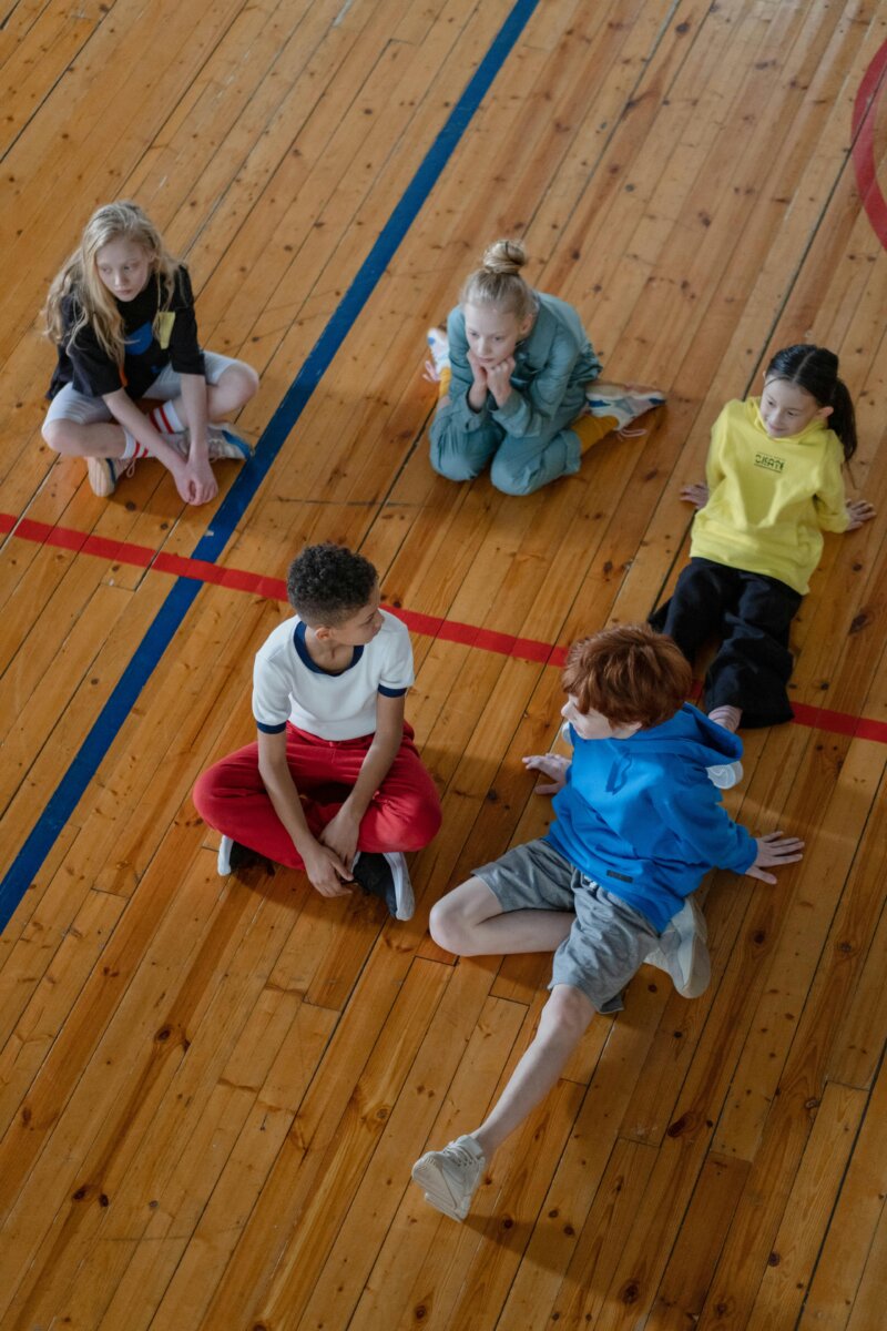 Group of diverse children sitting on gym floor, engaged in conversation, during physical education.